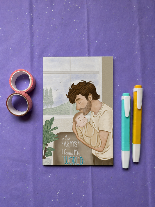 A square art print featuring an illustration of a father holding his baby with the text 'In Your Arms I Found My World', styled on a purple fabric background with washi tape and two pens.
